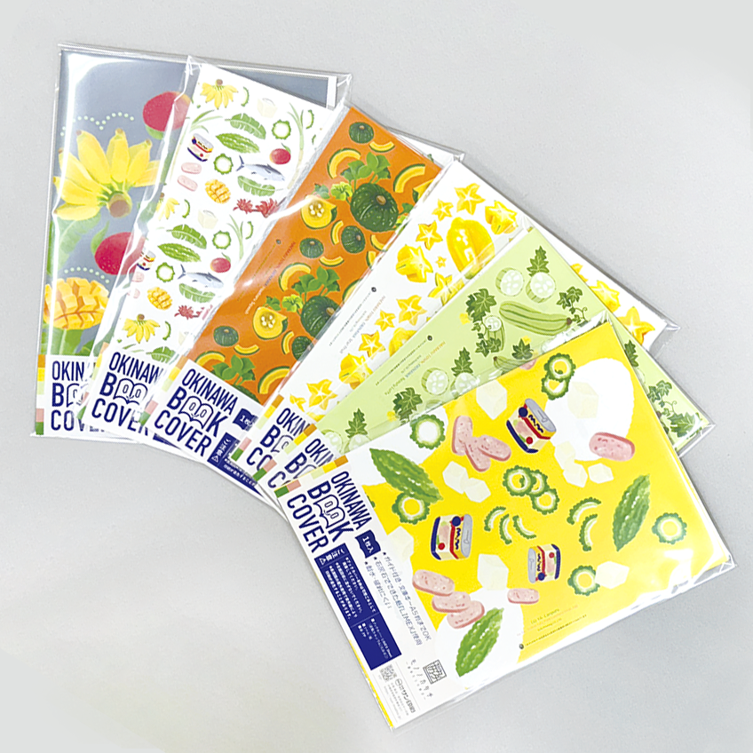 「オキナワブックカバー」ブックカバーが６枚並んでいる。ゴーヤーチャンプルー、食用ヘチマ、スターフルーツ、カボチャ、沖縄の食べ物や植物をコラージュしたもの、果物のデザイン。
